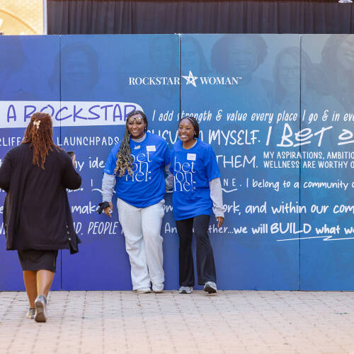 Two students in blue t-shirts stand against a backdrop created for a rockstar woman event on the Pew Grand Rapids campus while a person takes their photo